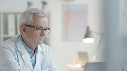 Tilt down shot of elderly male doctor in lab coat and glasses typing on laptop while working at desk in medical office