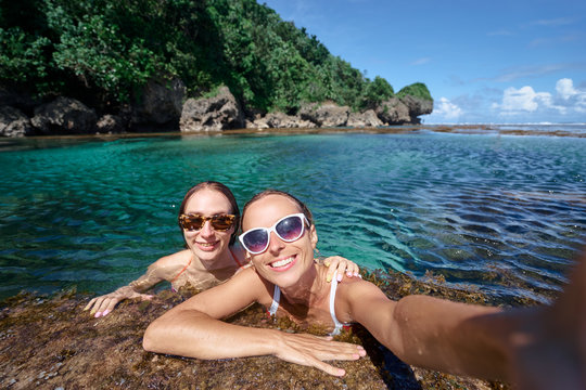 Vacation and technology. Colorful portrait of two pretty young women taking selfie portrait bathing in natural pool of tropical beach.