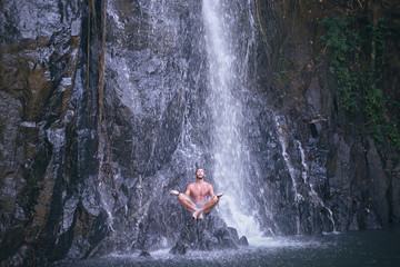 Fototapeta premium Wellness spa, vacation and yoga meditation concept. Young man sitting in lotus position on the rock under tropical waterfall.