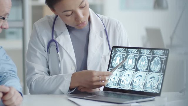 Tilt up shot of young mixed raced female doctor zooming and pointing at x-ray image of brain on laptop screen while explaining diagnosis to senior male patient during consultation