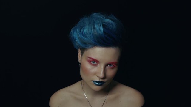 A girl with blue hair and make-up in red and blue colors posing in a dark studio. Fashion video shooting in blue and red lighting