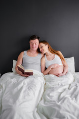 Happy family. Cozy home. Beautiful pregnant woman and her husband reading book together in bedroom.
