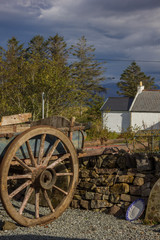 old wooden cart, large blade wheels, stone wall, white and blue plate on gravel, white houses with dark roof in the background and trees, cloudy sky