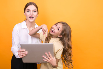 Beautiful woman with her happy daughter and laptop on orange background