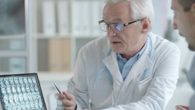 Elderly doctor showing x-ray image of brain on laptop and talking to male patient while giving consultation in medical office
