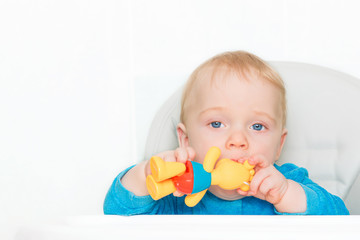 A kid with sad eyes holds his toy in his mouth. A blond boy is sitting in his chair with a toy in his hands on a white background. Close-up, empty space for text. The concept of a happy childhood.