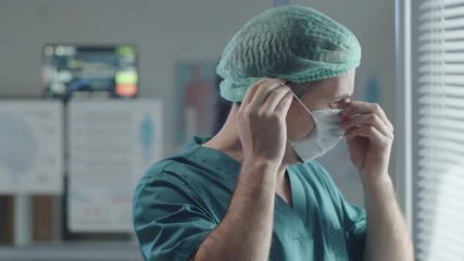 Tilt up shot of male hospital worker in scrubs and disposable hat putting medical mask on face and walking away - Powered by Adobe