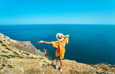 Back view of woman traveler in bright yellow dress and hat with backpack standing the top of mountain with raised hands against amazing seascape.