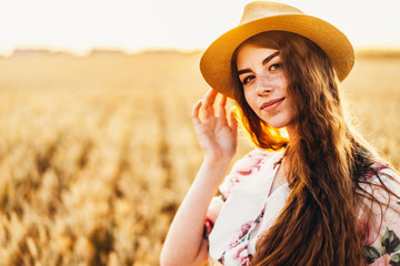 Portrait of a beautiful young woman with curly hair and freckles face. Woman in dress and hat posing in wheat field at sunset and looking at camera