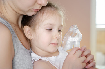  Cute little baby in the mask of an inhaler. Procedure of inhalation at home. Daughter and mother taking respiratory therapy with nebulizer.