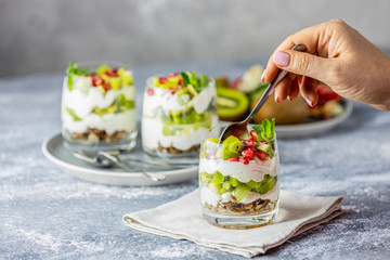 Person woman hand inserting spoon in the breakfast jar with granola, yogurt and kiwi fruits on gray surface. Delicious food, traditional american snack