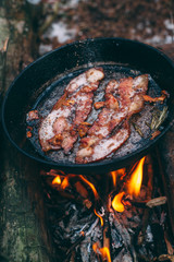 Slices of fried bacon in a pan. Food in a forest camp. Cooking on fire. Picnic in the nature. Grilled food on nature..