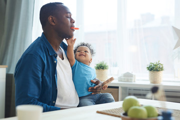 Portrait of happy African father playing with little son while sitting in sunlit kitchen, copy space