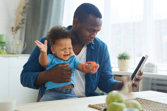 Side view portrait of African-American man holding crying baby while using digital tablet in home interior, copy space