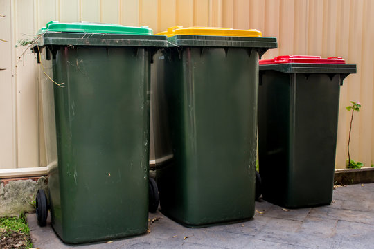 Australian garbage wheelie bins with red and yellow lids for general and recycling household waste and green lid for garden waste near the resedential building fence