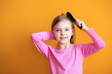 Cute baby, baby girl 6-8 years old combs her soft blond hair medium length comb and smiles on an orange background - Powered by Adobe