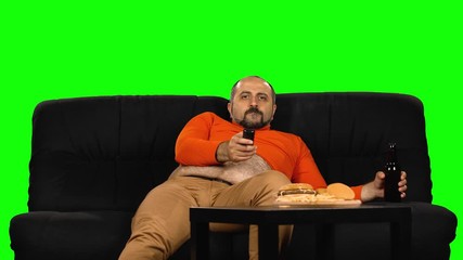 Caucasian man with big belly sits and laughs on black sofa near table with fast food, holds bottle of beer and TV remote. Green screen