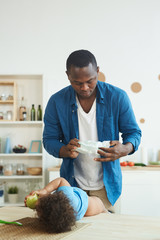 Vertical portrait of adult African-American man changing diaper while caring for toddler at home,...