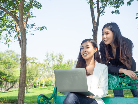 two asia business woman working with computer laptop in park. beauty woman tpying keyboard notebook blank screen and friend standing behind with copy space.  