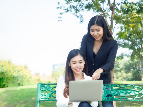 two asia business woman working with computer laptop in park. beauty woman tpying keyboard notebook blank screen and friend standing behind with copy space.  