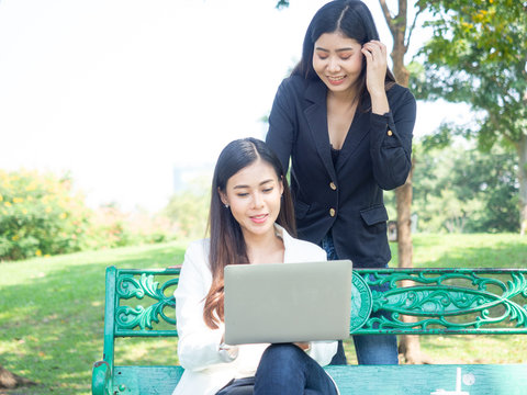 two asia business woman working with computer laptop in park. beauty woman tpying keyboard notebook blank screen and friend standing behind with copy space.  