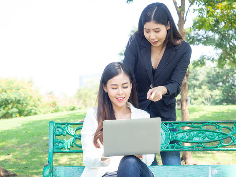 two asia business woman working with computer laptop in park. beauty woman tpying keyboard notebook blank screen and friend standing behind with copy space.  