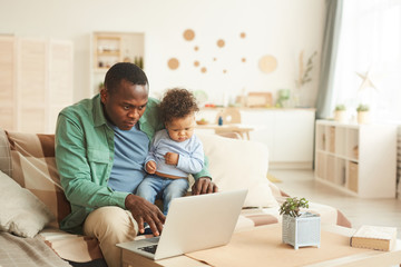 Portrait of mature African-American man working on laptop while babysitting son at home, copy space