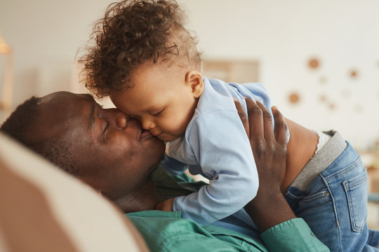 Side View Portrait Of Loving African-American Dad Kissing Baby Son While Playing At Home, Copy Space