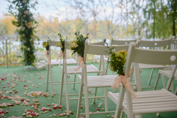 Fototapeta na wymiar White wood chairs with fresh flowers on each side of archway outdoods, copy space. Empty chairs prepared for wedding ceremony