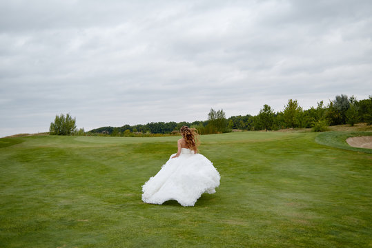 Portrait of beautiful bride in luxury white wedding dress with feathers running on green golf course, back view. Runaway bride, copy space