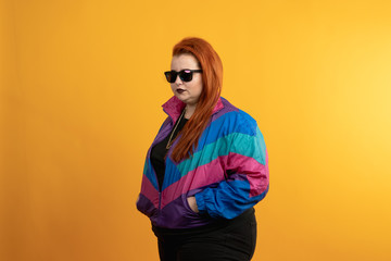 Overweight woman in sunglasses and windbreaker posing with hand in pocket isolated on yellow background © Ivan