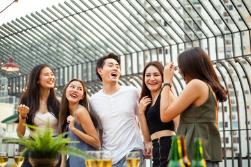 Group of happy Asian girls and man celebrating party, dancing and drink at rooftop in sunset together, female gang and boy friend having fun, chatting, laughing, night lifestyle of young people
