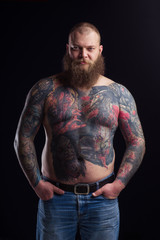 Tattooed brutal bearded man with a naked torso. Tattoo model portrait. Tattooing art artist. Brutal rocker biker stern big dork serious dangerous. Black background. Beard. 