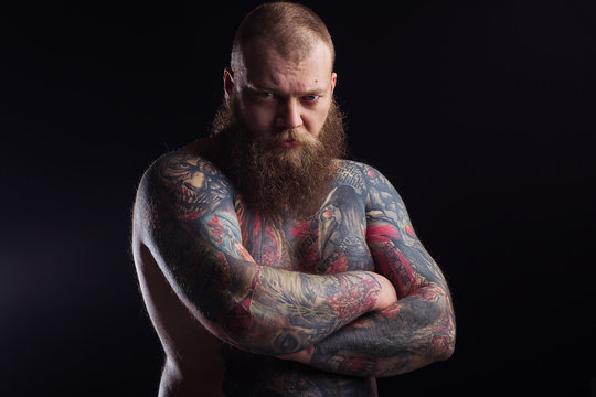 Tattooed brutal bearded man with a naked torso. Tattoo model portrait. Tattooing art artist. Brutal rocker biker stern big dork serious dangerous. Black background. Beard. 