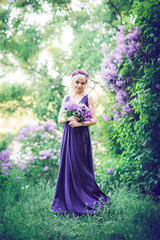 Obraz na płótnie Canvas Outdoor fashion photo of a beautiful young woman surrounded by purple flowers. Girls in a long dress with a slit on the background of a spring garden with lilacs. The concept of cosmetics and perfumes