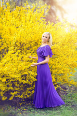 Fototapeta premium girl on a background of yellow flowers. Spring photo with a beautiful woman in a purple long dress with blooming forsythia. fabulous fantasy image . Spring holiday. Women's day
