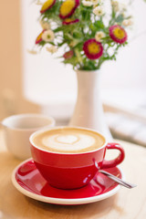 Red cup with hot milk coffee and vase with colorful flowers on a table.