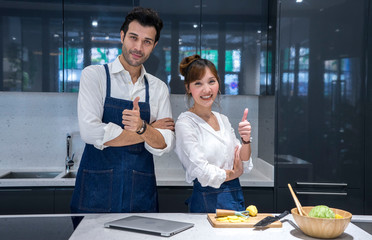 Asian woman couple with a handsome chef, thumbs up to cook healthy food, salads in the kitchen.