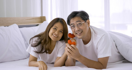 Happy asian couple holding red heart decorations together in a bedroom.