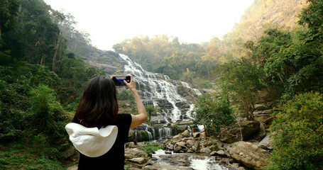Woman in black t-shirt taking photos of beautiful waterfall by smartphone.