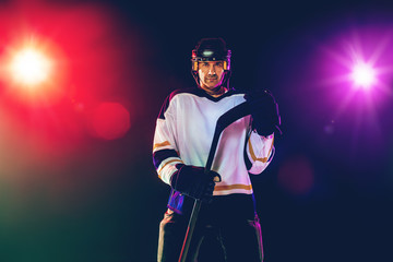 Fototapeta premium Winner. Male hockey player with the stick on ice court and dark neon colored background. Sportsman wearing equipment, helmet practicing. Concept of sport, healthy lifestyle, motion, wellness, action.