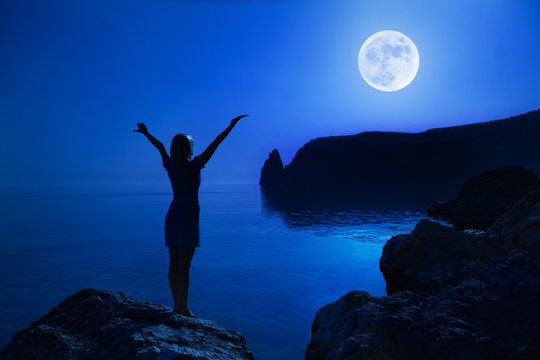 Rear view unidentified young happy woman stands on a stone raise Hands up Looking at the big moon and calm clear sea water against the backdrop of landscape and clear night sky. Full moon concept