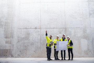 A group of engineers standing against concrete wall on construction site. - obrazy, fototapety, plakaty