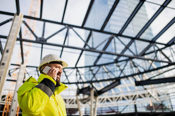 Man engineer standing on construction site, using smartphone.