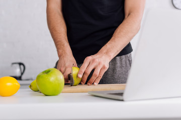 cropped view of man cutting apple near laptop at home