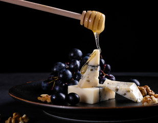 Peace of handmade cheese on plate with grapes, nuts and honey at black background. Close up.