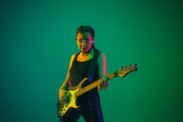 Rockstar. Caucasian female guitarist portrait on green studio background in neon light. Beautiful female model in black wear with guitar. Concept of human emotions, facial expression, ad, music, art.