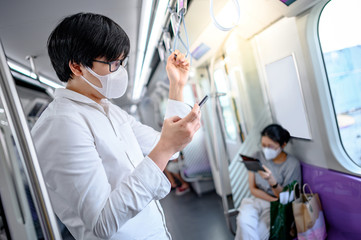 Asian man wearing surgical face mask using smartphone on skytrain or urban train. Wuhan coronavirus (COVID-19) outbreak prevention in public transportation. Health awareness for pandemic protection