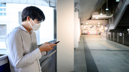 Naklejka na ściany i meble Asian man wearing surgical face mask using smartphone at skytrain station platform. Wuhan coronavirus (COVID-19) outbreak prevention in public transportation. Health awareness for pandemic protection