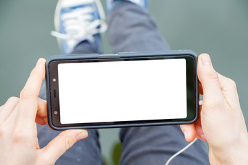the girl holds a mobile phone in her hands and looks at its screen, the headphone cord is visible, in the background the girl's legs in jeans and sneakers, there is an empty space